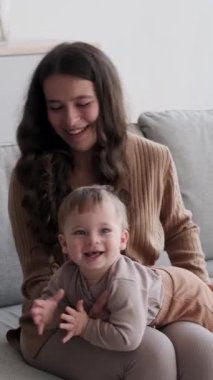 Kafkasyalı mutlu anne ve küçük oğlu evde oturma odasında birlikte oynuyorlar, kanepede oturuyorlar. Annelik, çocukluk. Dikey video.