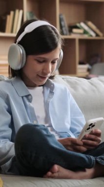 Neşeli beyaz çocuk internette geziniyor ve telefonla müzik dinliyor, hafta sonu evde boş vakitlerinde kanepede dinleniyor. Dikey video.