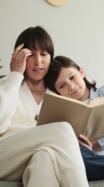 Arkadaş canlısı Kafkas anne ve okul çocuğu birlikte kitap okuyor, evde boş vakitlerinde kanepede dinleniyorlar. Aile bağı ve boş zaman. Dikey video.