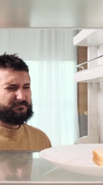 Mutsuz ve mutsuz orta yaşlı beyaz adam akşamdan kalma ve boş buzdolabında kuru veya bayat sandviç yiyor. Dikey video.