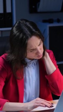 Çok çalışan ve yorgun orta yaşlı beyaz iş kadını gece geç saatlerde çalışırken boynu ağrıyor. Dikey video.