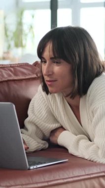 Gülümseyen beyaz kadın internette dizüstü bilgisayarla geziniyor, evdeki kanepede uzanıyor ve internette zaman geçiriyor. Dikey video.