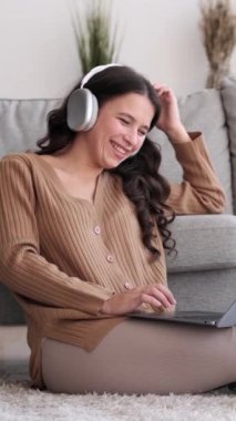 Dost canlısı, kulaklık takan beyaz bir kadın internette boş vakitlerini dizüstü bilgisayarla geçiriyor ve evde gülüyor. Dikey video.