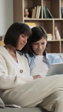 Mutlu Kafkasyalı orta yaşlı anne ve genç oğlu online eğlence ve kahkaha için tablet kullanıyor. Evdeki kanepede dinlenirken. Dikey video.