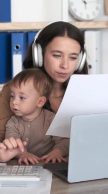 Evdeki ofiste dizüstü bilgisayarla çalışan neşeli bir kadın ve yeni yürümeye başlayan bir oğlu var. İnternette çalışmak ya da çocukla internet öğrenmek. Dikey video.