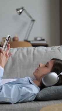 Cep telefonlu, kanepeye uzanmış, evdeki oturma odasında interneti karıştıran sıkıcı beyaz bir çocuk. Çevrimiçi boş zaman kavramı. Dikey video.