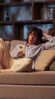 Uykucu Kafkasyalı orta yaşlı kadın cep telefonunu sosyal medyada mesajlaşmak için kullanıyor ve esniyor, evdeki kanepede uzanıyor. Dikey video.