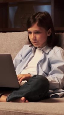 Neşeli Kafkasyalı genç öğrenci akşam evde dizüstü bilgisayarla vakit geçiriyor. Online eğlence kavramı. Dikey video.