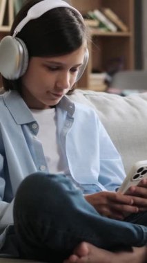 Evde telefon mesajları ve internet tarayıcıları kullanan mutlu beyaz bir çocuk. Boş zamanlarını müzik dinleyerek geçiriyorsun. Dikey video.