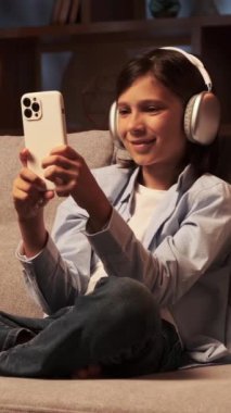 Neşeli Kafkasyalı genç okul çocuğu internet eğlencesi için akıllı telefon kullanıyor ve akşam evde kulaklıkla müzik dinliyor. Dikey video.