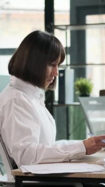 Modern ofisten online çalışırken evrak işleri ve iş muhasebeleriyle uğraşan ciddi bir beyaz kadın serbest çalışan. Dikey video.