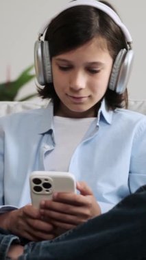 Neşeli, beyaz, kulaklık takan, müzik dinleyen ve sosyal medyada akıllı telefon kullanarak mesajlaşan bir öğrenci. Dikey video.