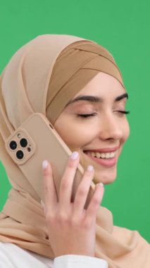 Olumlu Arap genç kadın, yeşil arka plandaki arkadaşlarını aramak için akıllı telefonu kullanıyor. Uzaktan konuşma kavramı. Dikey video.