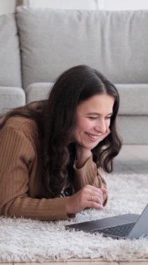 Neşeli beyaz kadın internette geziniyor, sosyal medyada mesajlaşıyor ve evinde dizüstü bilgisayar kullanarak gülüyor. Dikey video.