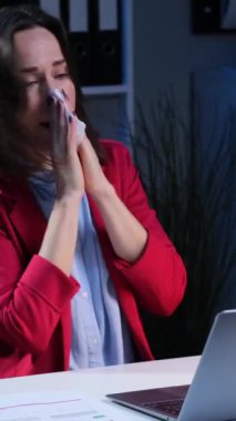 Sağlıksız ve üzgün orta yaşlı beyaz iş kadını akşam ofisinde dizüstü bilgisayarla çalışırken peçeteye hapşırıyor. Dikey video.
