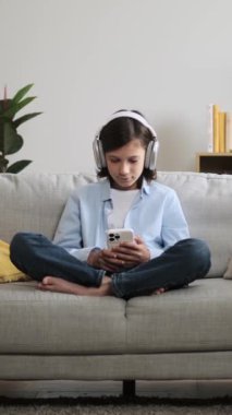 Beyaz bir çocuğun evde oturma odasındaki kanepede dinlenirken akıllı telefonuyla sohbet etmesi ve kulaklıkla müzik dinlemesi. Dikey video.