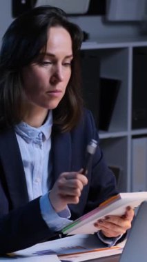 Düşünceli, beyaz, orta yaşlı bir çalışan gece geç saatlerde ofiste çalışırken not defterine veya takvime not alıyor. Dikey video.