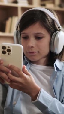 Kulaklık takan, interneti gezen, boş zaman geçiren ve evde gülen mutlu beyaz bir çocuk. Dikey video.
