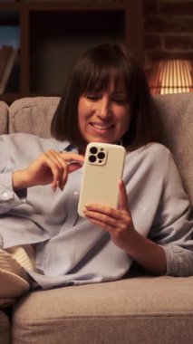 Kafkasyalı neşeli orta yaşlı kadın kanepeye uzanıyor, telefon harcamak için zaman harcayıp internette mesajlaşıyor ve akşam evde gülüyor. Dikey video.