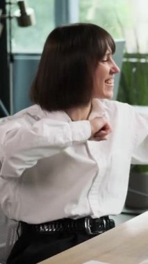 Orta yaşlı mutlu beyaz kadın dans ediyor ve ofiste çalışırken başarı ya da başarıyı kutluyor. Dikey video.