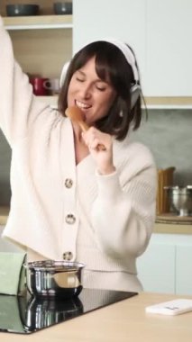 Gülümseyen orta yaşlı beyaz kadın mutfakta yemek pişiriyor, kulaklıkla müzik dinliyor ve kaşık kullanarak şarkı söylüyor. Dikey video.