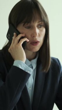 Ofiste çalışırken müşteriyle konuşmak için cep telefonu kullanan neşeli beyaz, orta yaşlı bir iş kadını. Dikey video.