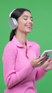 Neşeli kadın cep telefonuyla eğleniyor ve gülüyor, yeşil arka planda duruyor. Dikey video.