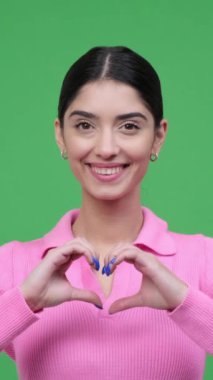 Gülümseyen Kafkasyalı genç kadın yeşil arka planda kalp şeklinde jest yapıyor. Aşkın, sevginin ve romantizmin sembolü. Dikey video.