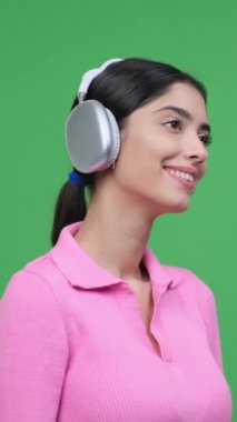 Arkadaş canlısı Kafkas genç kadın kablosuz kulaklıkla müzik dinliyor ve eğleniyor, yeşil arka planda boş zamanlarında dinleniyor. Dikey video.