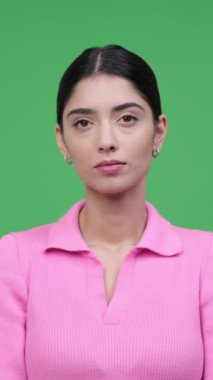Ciddi ve gururlu beyaz bir genç kadının portresi kameraya bakıyor ve yeşil arka planda duruyor. Konsantre ve kendinden emin bayan. Dikey video.