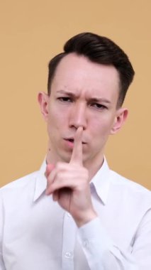 Konsantre ve odaklanmış beyaz bir adam bej arka plan hakkında gizli bilgi tutmak için sessiz jest yapıyor. Dikey video.