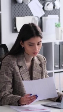 Laptop ve belgelerle çalışan düşünceli, beyaz, genç bir kadın ofiste masa başında oturuyor. Strateji geliştiriyorum. Dikey video.