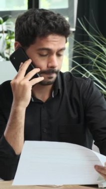Belgelerle uğraşan konsantre Ortadoğulu bir adam ve ofiste müşterileriyle sohbet ediyor. Dikey video.