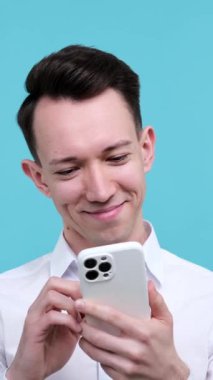 Sosyal medyayı karıştıran ya da akıllı telefon kullanarak mesajlaşan, mavi arka planda duran Kafkasyalı genç bir adam. Dikey video.