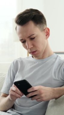 Akıllı telefonlu, evdeki koltukta internete girip siber alemde boş zaman geçiren düşünceli, beyaz bir genç adam. Dikey video.