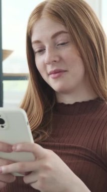 Beyaz kız akıllı telefonu kullanarak online sosyal medyada ve evde internet eğlencesinde mesaj atıyor. Dikey video.