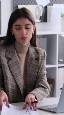 Ofiste kağıtlarla uğraşırken bilgisayarla iş arkadaşlarıyla çevrimiçi sohbet eden kızgın beyaz kadın. Dikey video.