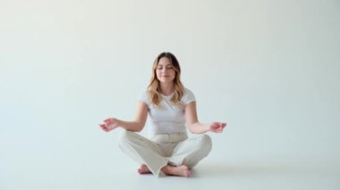 Genç memnun ve gülümseyen beyaz gözlü beyaz kadın, beyaz arka planda nilüfer çiçeğiyle yoga yapıyor ve meditasyon yapıyor. Ayrılma ve zihinsel denge kavramı.