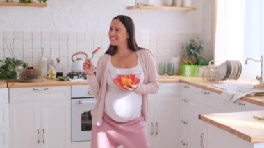 Gülümseyen ve hamile orta yaşlı beyaz kadın sağlıklı yiyeceklerin ve mutfakta müzik eşliğinde dans etmenin tadını çıkarıyor. Mutlu bebek beklentisi konsepti.