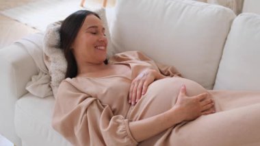 Neşeli, beyaz, orta yaşlı bir kadın. Kanepede dinleniyor ve karnına dokunuyor. Hamilelik kavramı, sevgi ve beklenti..