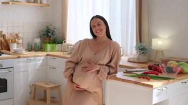 Dost canlısı, orta yaşlı, hamile bir kadının portresi karnına dokunuyor ve ev mutfağındaki kameraya bakıyor. Çocuk beklentisi kavramı.