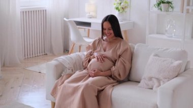 Hamile, orta yaşlı, beyaz bir kadın. Oturma odasındaki koltukta oturan hamile karnına nazikçe dokunuyor. Hamilelik, annelik ve rahatlama..