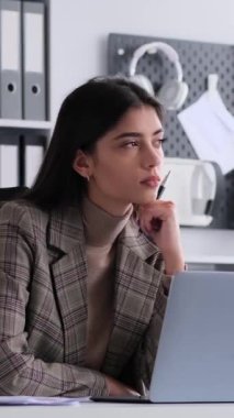 Düşünceli, genç, beyaz bir kadın laptopla düşünüyor, çalışıyor, muhasebe işleriyle uğraşıyor ve ofiste mali planlar yapıyor. Dikey video.
