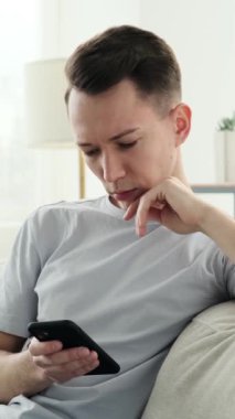 Neşeli, genç, beyaz bir adam internette akıllı telefon kullanarak eğleniyor ve evde gülüyor. Online eğlence kavramı. Dikey video.