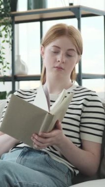 Kafkasyalı ciddi bir genç kız kitap okuyor, evinde kanepede oturuyor. Aile içi rahatlama ve edebiyat kavramı. Dikey video.