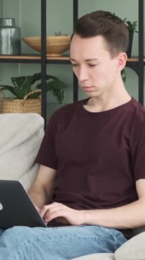 Dost canlısı, genç, beyaz bir adam internet eğlencesi için dizüstü bilgisayar kullanıyor ya da internette çalışıyor, evdeki kanepede dinleniyor. Dikey video.