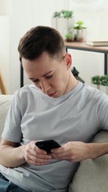 Cep telefonlu, internetten internete girip gülen ve evdeki kanepede dinlenen mutlu, genç beyaz bir adam. Dikey video.