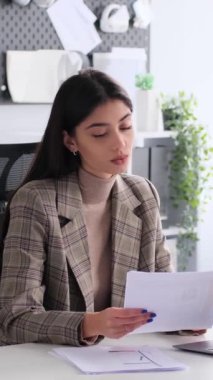 Ofiste doküman ve dizüstü bilgisayarla çalışırken çevrimiçi video görüşmesi yapan Kafkasyalı bir iş kadını. Dikey video.