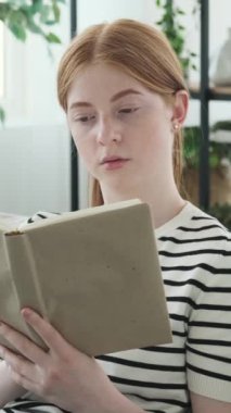 Keyifli Kafkasyalı kızıl saçlı kız evde boş zaman geçirirken kitap okuyor. Edebiyat hobisi kavramı. Dikey video.