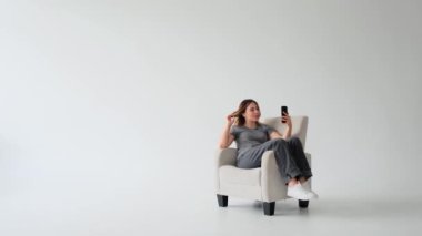 Olumlu, genç, beyaz bir kadın koltukta dinleniyor ve beyaz arka planda cep telefonuyla görüntülü konuşuyor. Dijital sohbete ve iletişime daldı.
